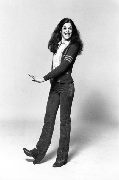 Gilda Radner poses in jeans and a cardigan sweater