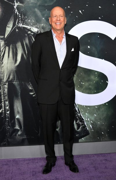 Bruce Willis stands with his hands behind his back in a black suit