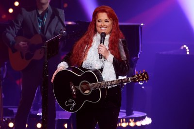 Wynonna Judd holds microphone and guitar while wearing a white shirt and black blazer 