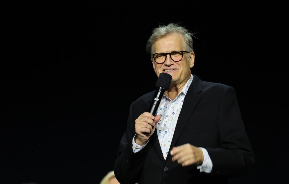 Drew Carey holds microphone up to his mouth while on stage in a black suit