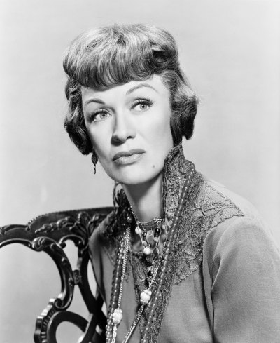 Eve Arden sits in chair and poses in a lace-neck top with necklaces