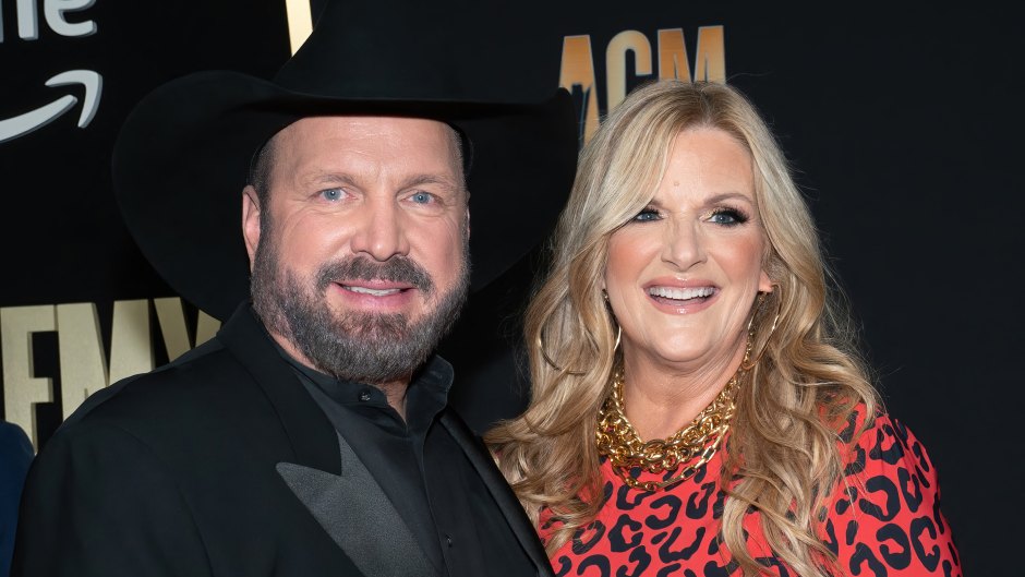 Garth Brooks, wearing an all black suit, poses next to wife Trisha Yearwood who's wearing a red leopard print dress.