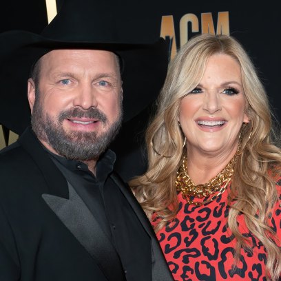 Garth Brooks, wearing an all black suit, poses next to wife Trisha Yearwood who's wearing a red leopard print dress.