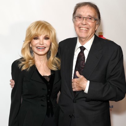 Loni Anderson smiles while wearing a black blazer and skirt next to husband Bob Flick