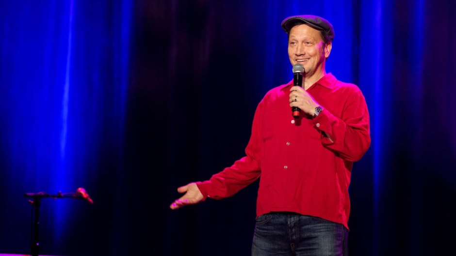 Rob Schneider performs on stage in a red sweater