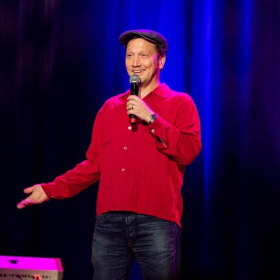 Rob Schneider performs on stage in a red sweater