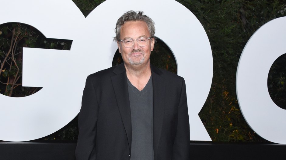 Matthew Perry smiles at camera in a black suit with a gray shirt