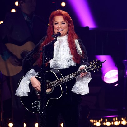 Wynonna Judd performs with a guitar on stage in a black blazer and white shirt