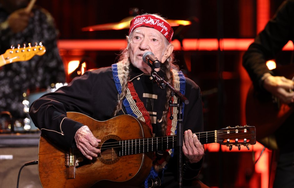 Willie Nelson holds a guitar on stage while wearing a red bandana