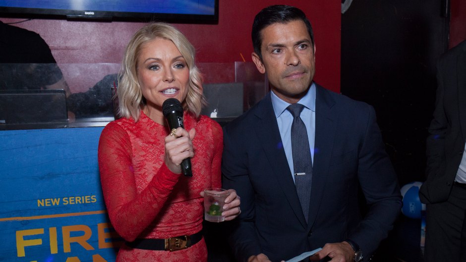 Kelly Ripa talks into a microphone into a red dress next to Mark Consuelos