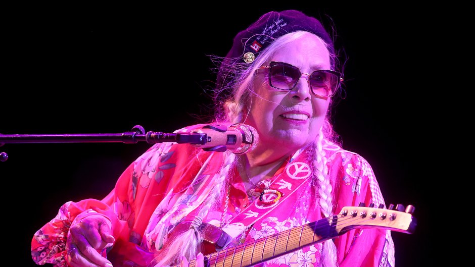 Joni Mitchell performing on stage with a guitar while wearing a pink shirt and sunglasses.