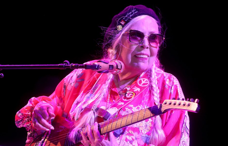 Joni Mitchell performing on stage with a guitar while wearing a pink shirt and sunglasses.